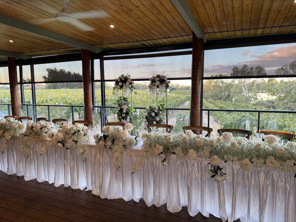 A long head table dressed in white with a full front of white flowers and two tall flower stands behind it