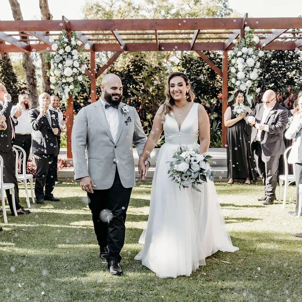 A couple walking back down the aisle under a pergola dressed with white flowers