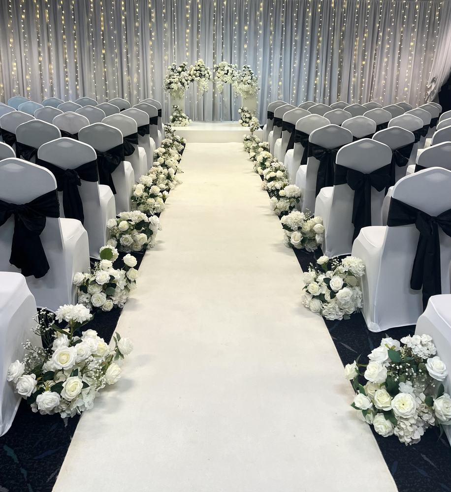 An indoor aisle lined with white flower arrangements, chairs with dark sashes and a flower arch at the end