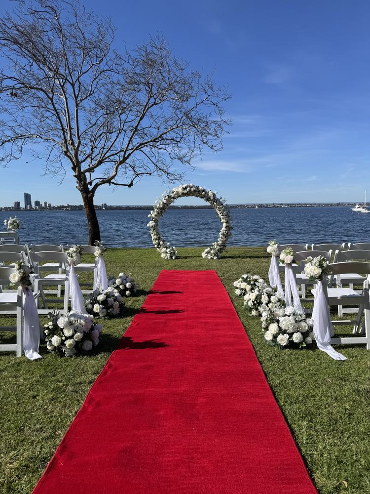 An outdoor ceremony by the river with a round floral arch, white flower clusters along a red aisle and chairs dressed with white fabric