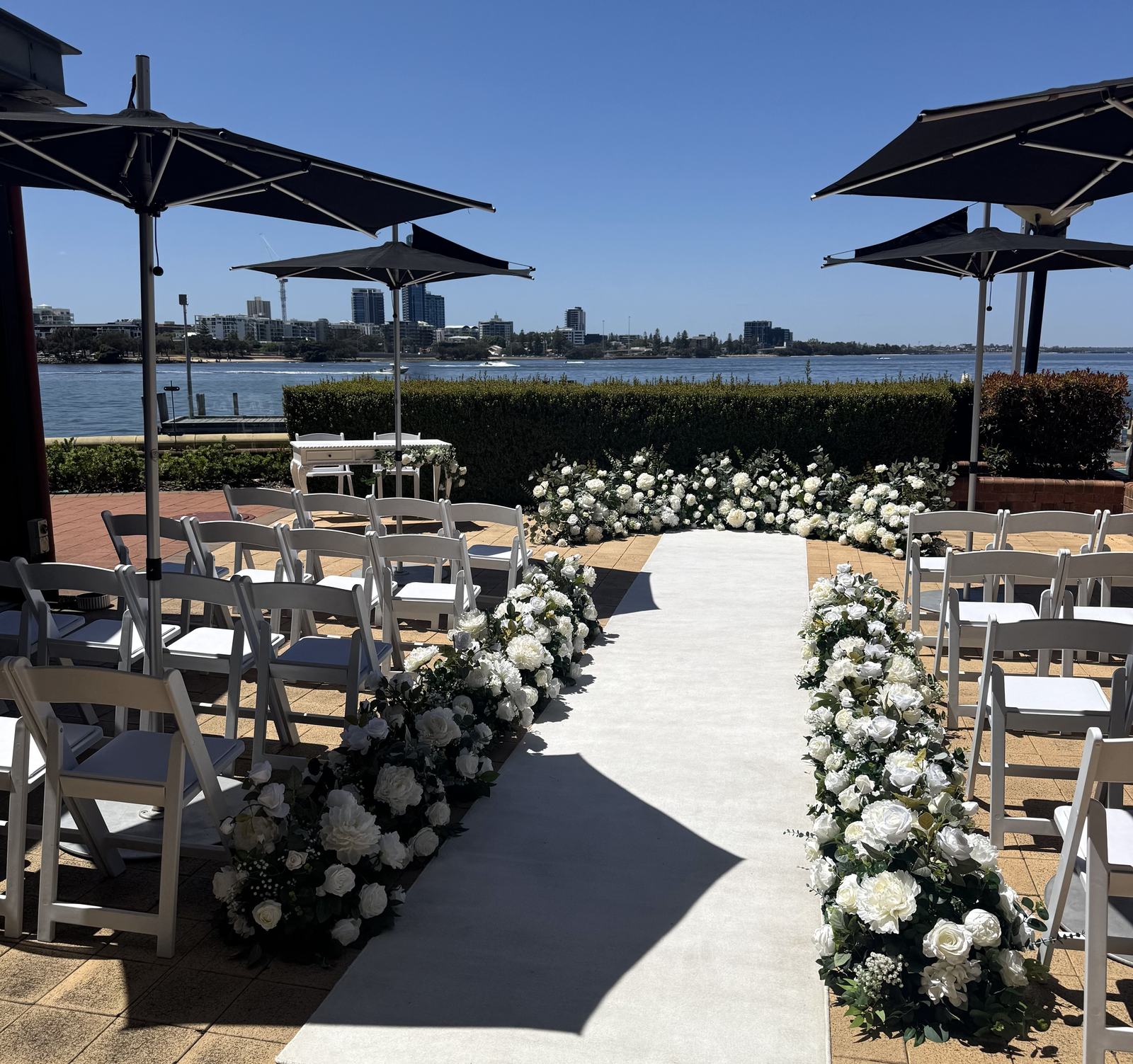 A waterside ceremony set-up with white flower arrangements lining a white aisle and dark carpet, umbrellas and the river behind