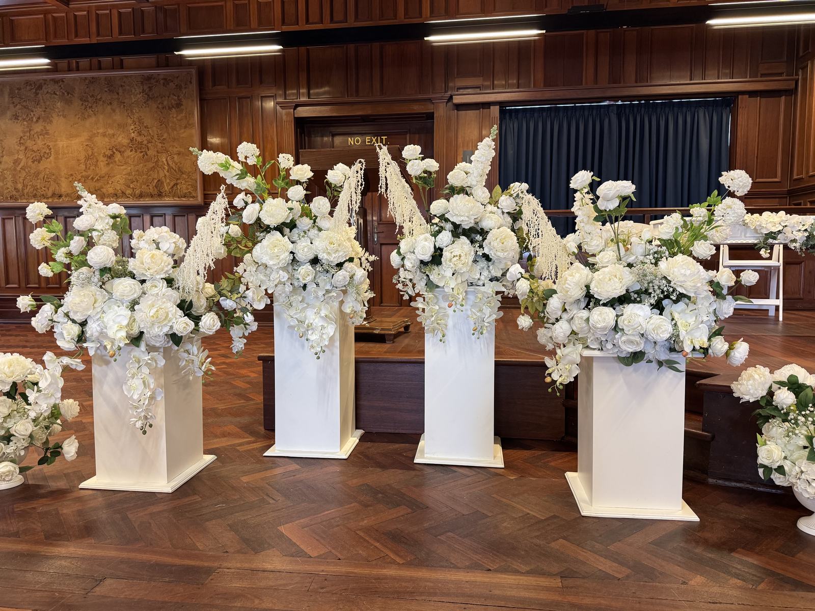 Four white pedestals topped with tall arrangements of white flowers in a wood-panelled room