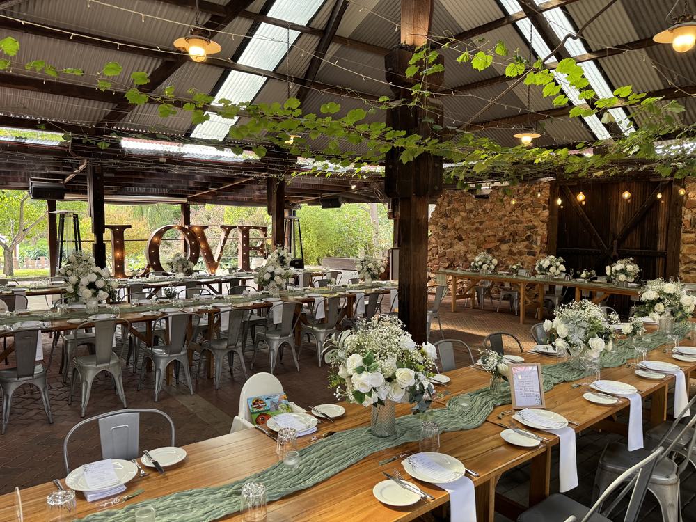 Long guest tables with green runners and white flower centrepieces under a vine-covered roof